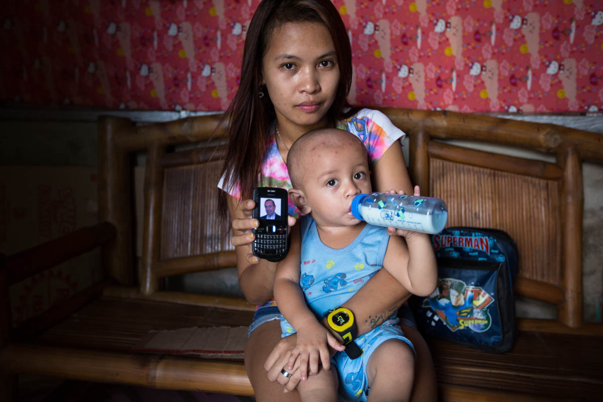 Joylyn and Renzo - Joylyn, 21 and Renzo, 1, in their home showing the only photo of Renzo’s, father, an Australian citizen on a cell phone. Joylyn and Renzo, get no support from him and have lost his contact.  Angeles City, Philippines, June 2015. - Copyright © © S. Borcard - N. Metraux - Angeles - Central Luzon - Philippines - <A href="https://maps.google.com/?ll=15.17343,120.601083&z=16" target="_blank">(Map Joylyn and Renzo)</A>